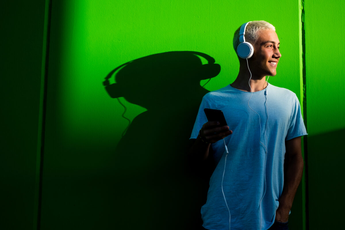 Un adolescent portant des écouteurs écoute un podcast. Derrière lui, son ombre sur mur vert sous-entend qu'il écoute un balado héberger sur Spotify